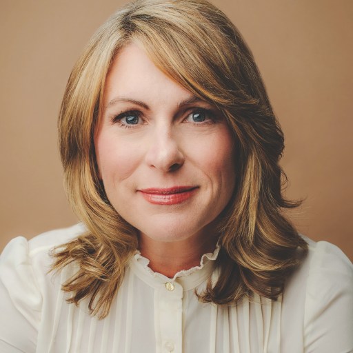 A person with shoulder-length blonde hair and a white blouse smiles slightly against a neutral background.