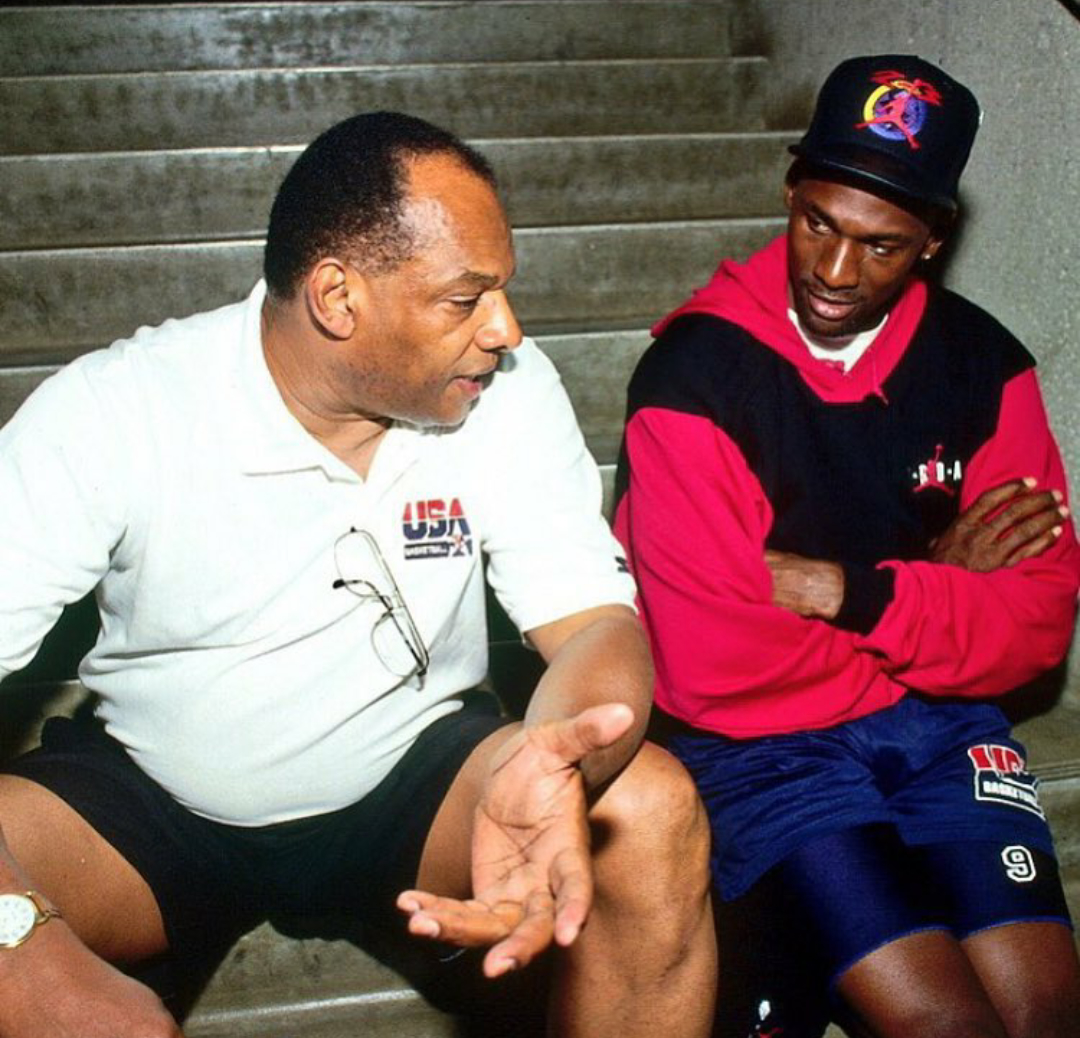 Two men seated on stairs engaged in conversation. The man on the left wears a white polo, and the man on the right wears a cap and a USA-branded sports outfit.