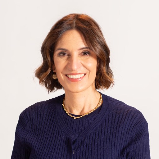 A woman with shoulder-length brown hair smiles, wearing a navy sweater and gold necklace, set against a plain background.