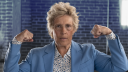 Person wearing a light blue suit and floral shirt, flexing both arms, set against a brick wall background.