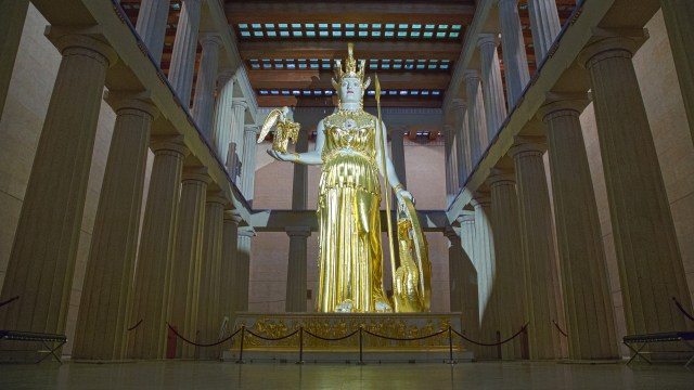 The golden statue of Athena stands prominently in a replica of the Parthenon interior, steeped in mythology. She holds a shield and a small figure, while the spacious setting boasts large columns and high ceilings.