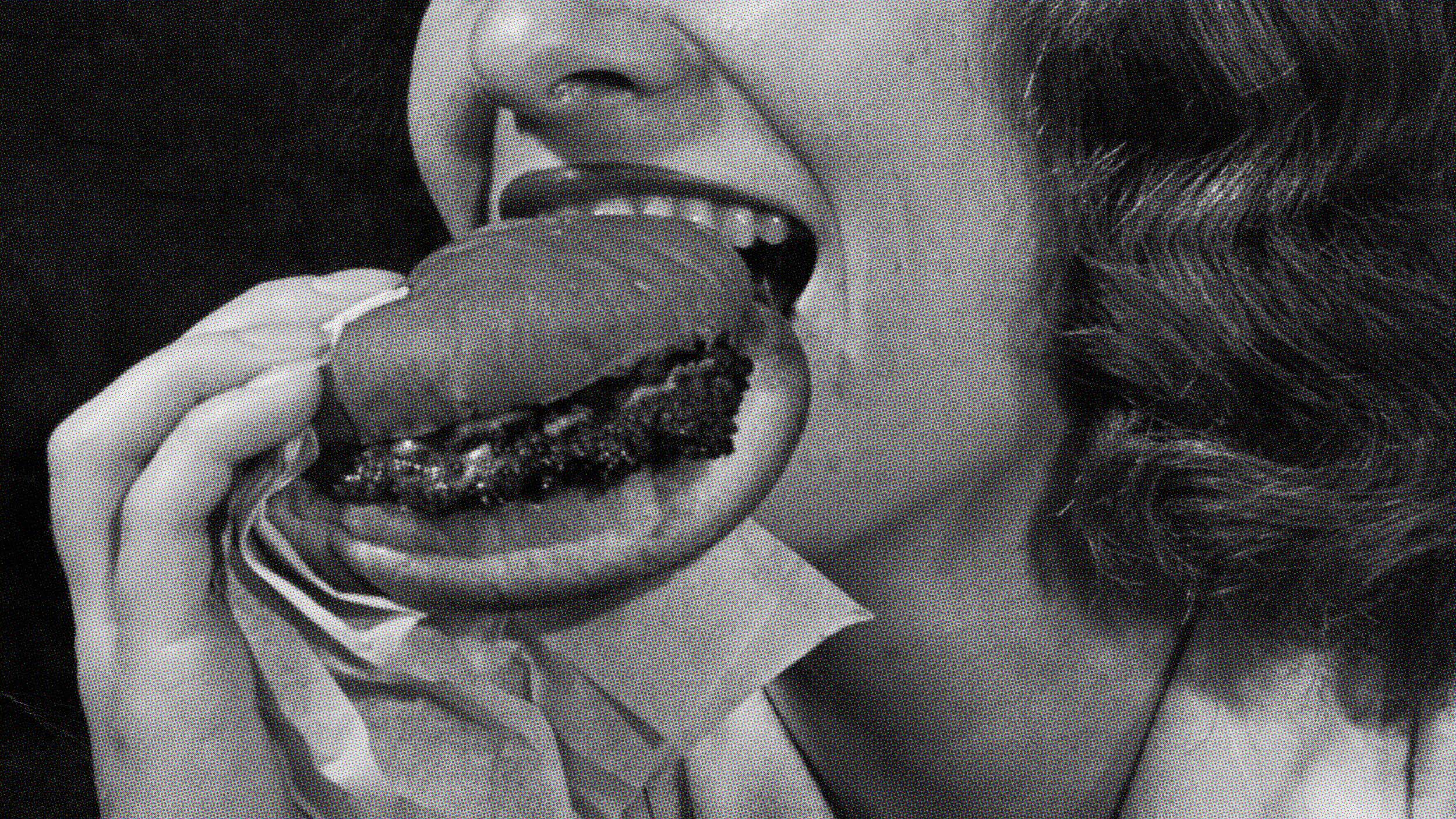 Black and white close-up of a woman savoring a burger wrapped in paper, her eyes reflecting her elevated taste expectations.