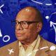 Man in glasses and a brown jacket with a serious expression; the background features a blue overlay adorned with white sports strategy symbols and a cheering crowd. It's reminiscent of a Coach Raveling masterclass, where every move is meticulously planned.