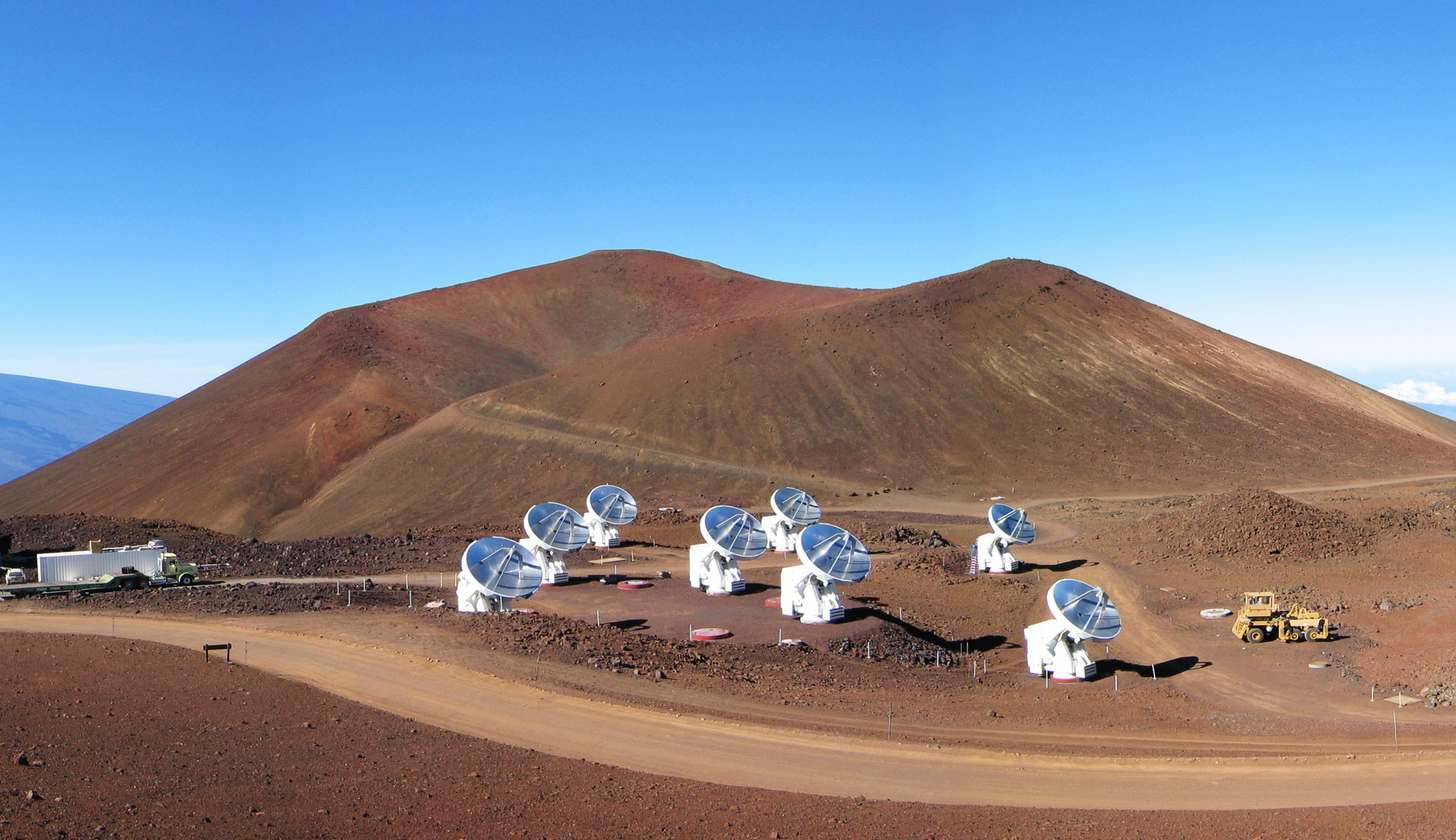 Array of large satellite dishes on a barren, reddish landscape beneath a clear blue sky, with a dirt road curving in front of them. As the hills stand sentinel in the background, these instruments seek to unravel cosmic mysteries like the ring nebula's true structure.