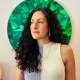 A woman with long curly hair stands in front of a green geometric background, wearing a sleeveless two-tone top and a necklace.