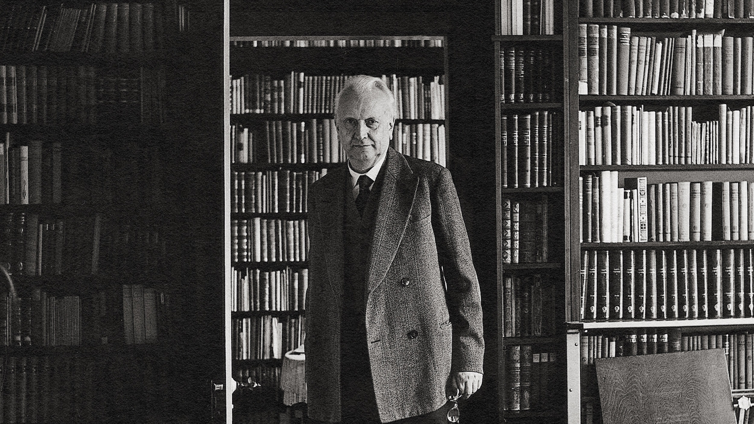 An older man in a suit stands in front of a large bookcase filled with books, pondering life's limit situations.