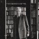 An older man in a suit stands in front of a large bookcase filled with books, pondering life's limit situations.