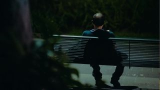 A lifelong single with short hair sits alone on a park bench at night, facing away. Bushes and dim lighting surround the scene.