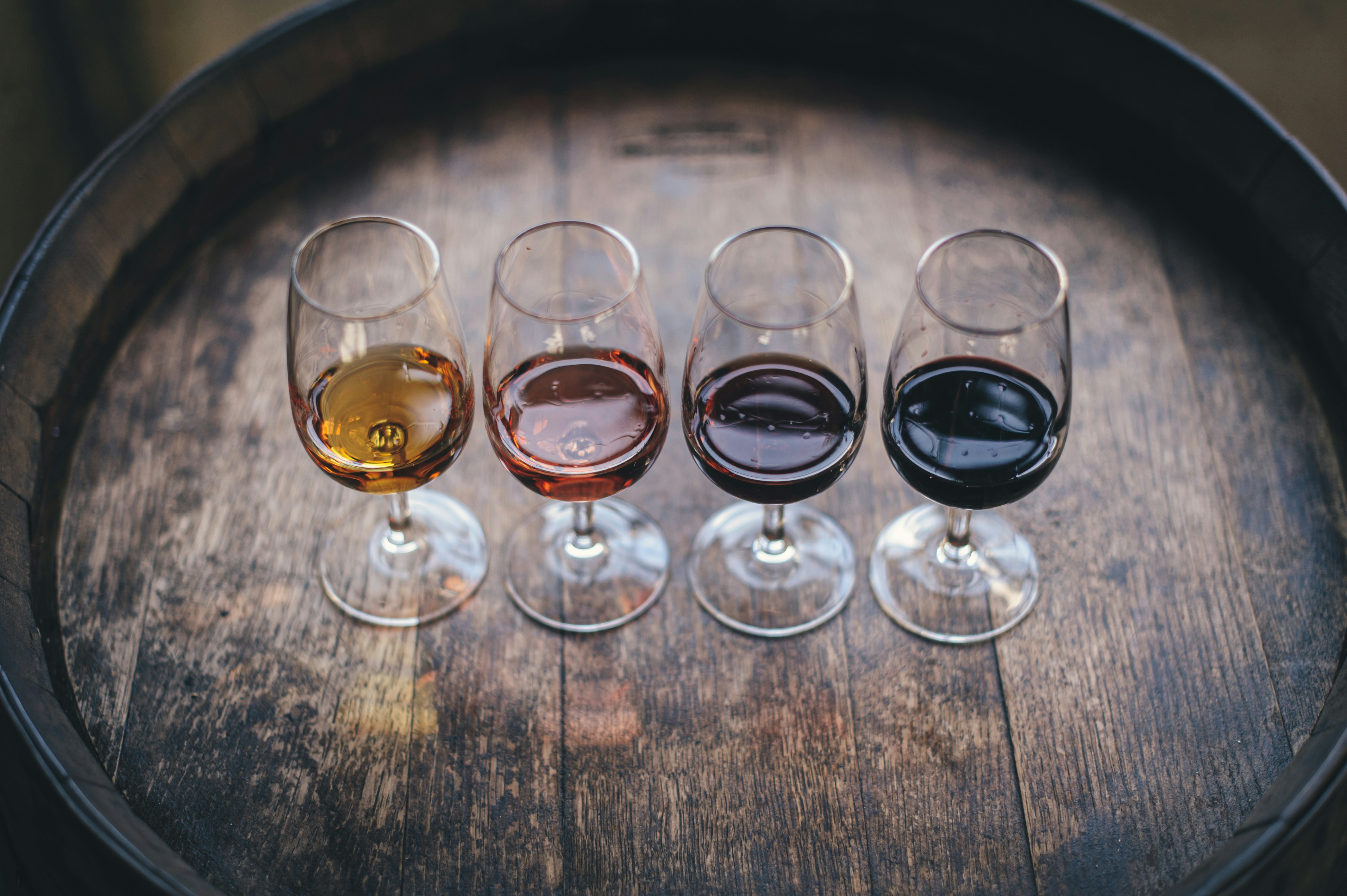 Four glasses of wine in varying shades from light to dark are arranged in a row on a wooden barrel.