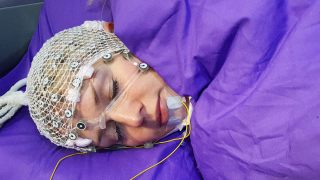 Person sleeping with EEG cap and sensors on face, covered with a purple blanket.