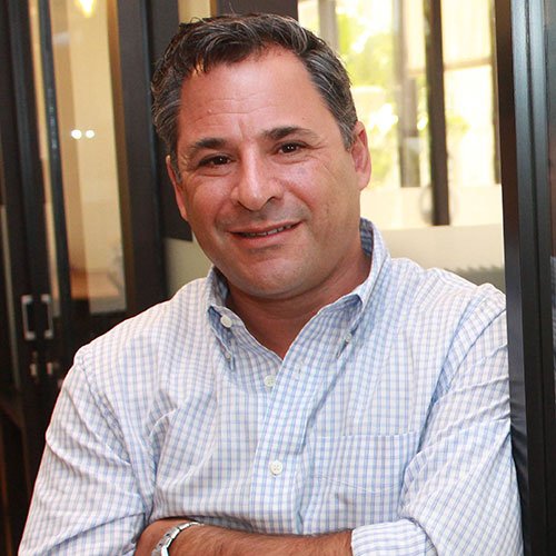 A man in a checkered shirt stands with his arms crossed, smiling, in an office setting with glass walls.