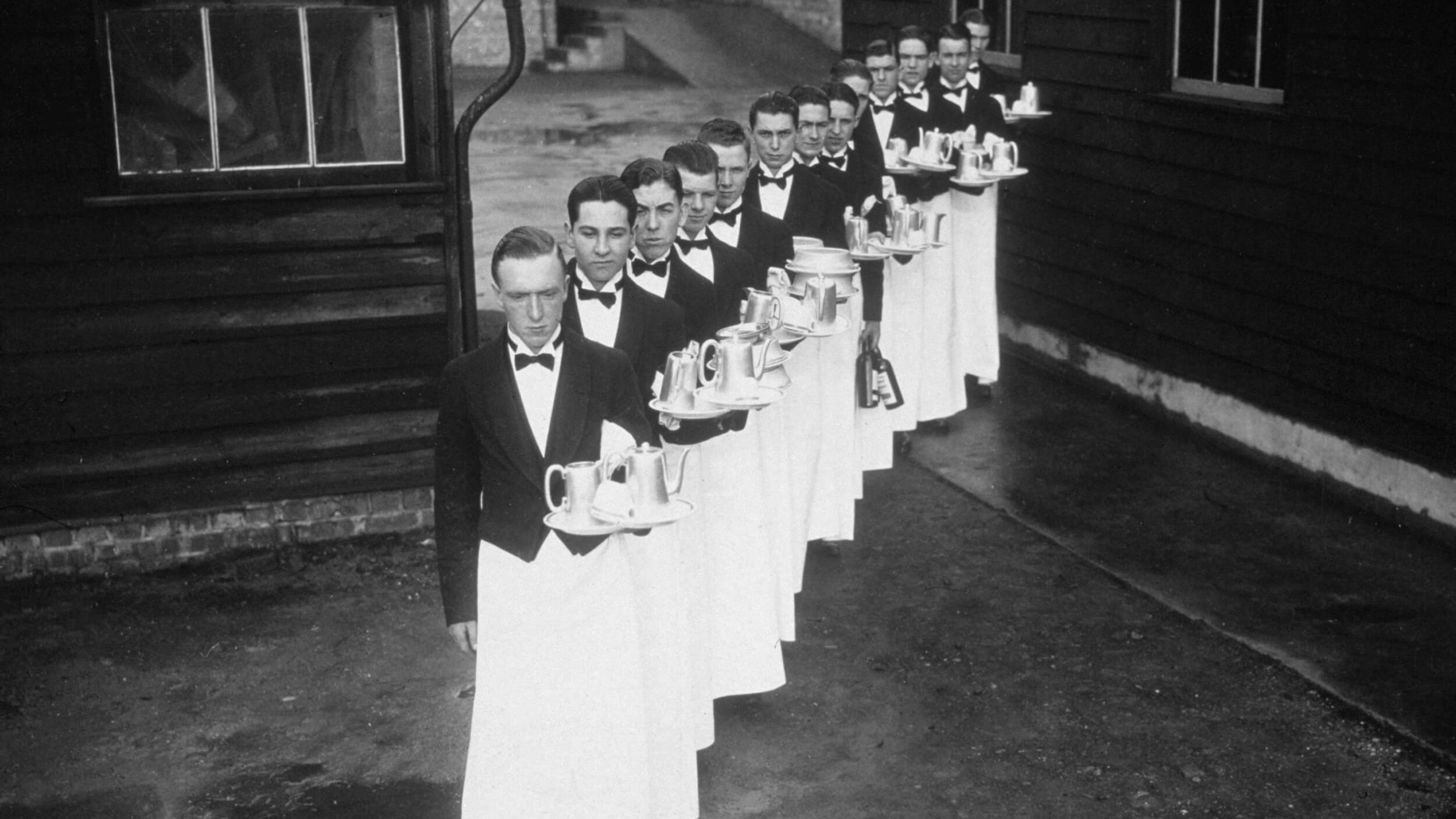 A line of waiters in formal attire, embodying a sense of unreasonable hospitality, carry trays with teapots and cups as they gracefully walk in procession outside the building.