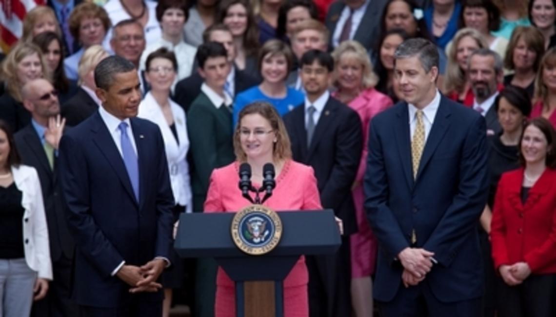 Amid a large outdoor gathering, a woman speaks at a podium adorned with a presidential seal, addressing the pressing issue of science illiteracy. Flanked by two men in suits, she emphasizes the importance of bridging gaps in knowledge to combat widespread ignorance.