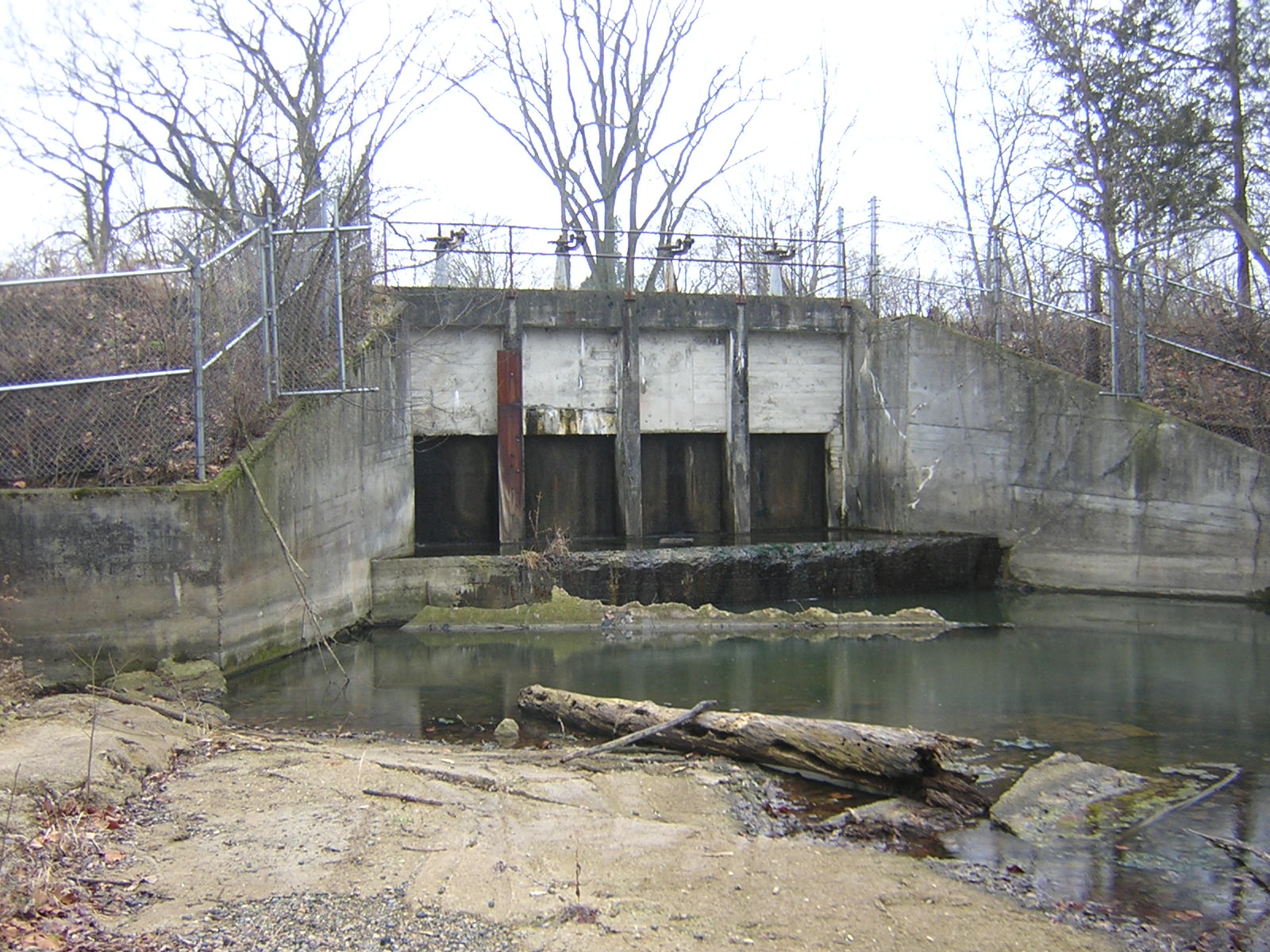 Concrete dam with three closed gates, surrounded by a chain-link fence. Logs and debris are visible in the water below. Leafless trees in the background.