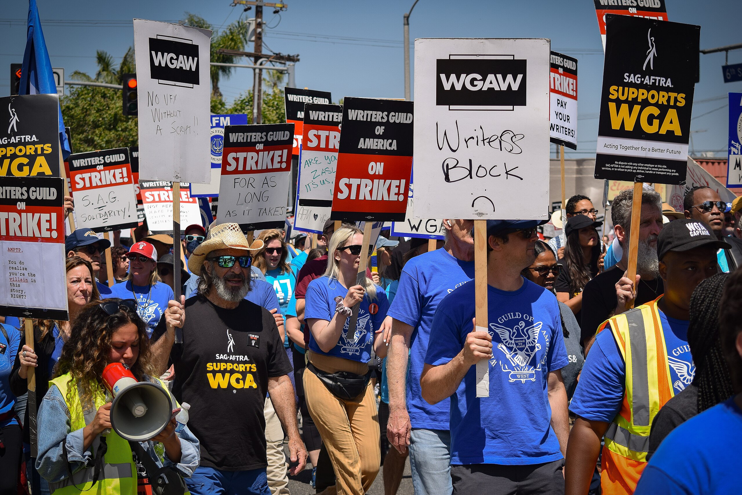 Protestors holding signs, such as "Writers Guild on Strike" and "Writers Block," gather in a street demonstration, challenging the rise of reality TV as they strive for fair compensation and creative rights.