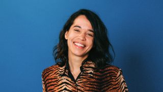 A person with long dark hair beams with creativity, wearing a tiger print jacket, set against a vibrant blue background.