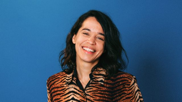 A person with long dark hair beams with creativity, wearing a tiger print jacket, set against a vibrant blue background.