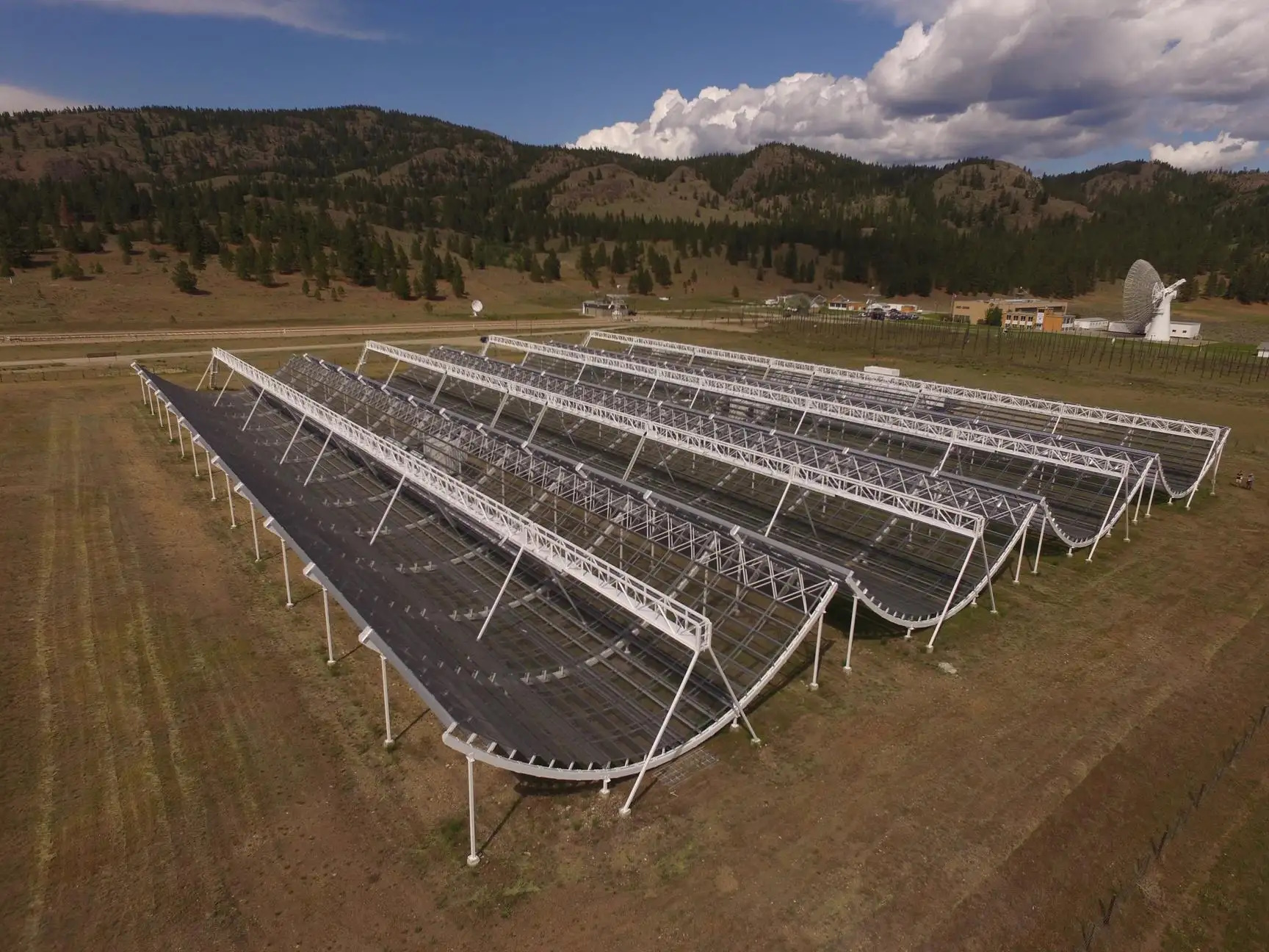 A large radio telescope array, reminiscent of LIGO's vital role in gravitational wave detection, features multiple curved antennas on a grassy plain, surrounded by hills and scattered buildings under a partly cloudy sky.