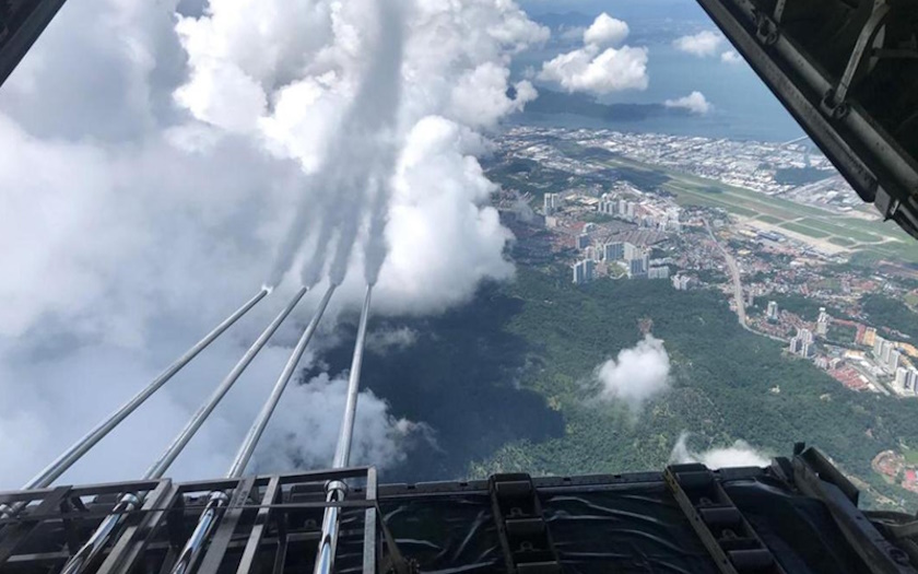 Aircraft releases cloud-seeding agents into the sky above a cityscape and forest, viewed from the open rear of the plane.