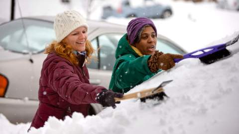 Two people experience everyday enlightenment as they shovel snow off a car in a winter wonderland, bundled up in cozy jackets, hats, and gloves.