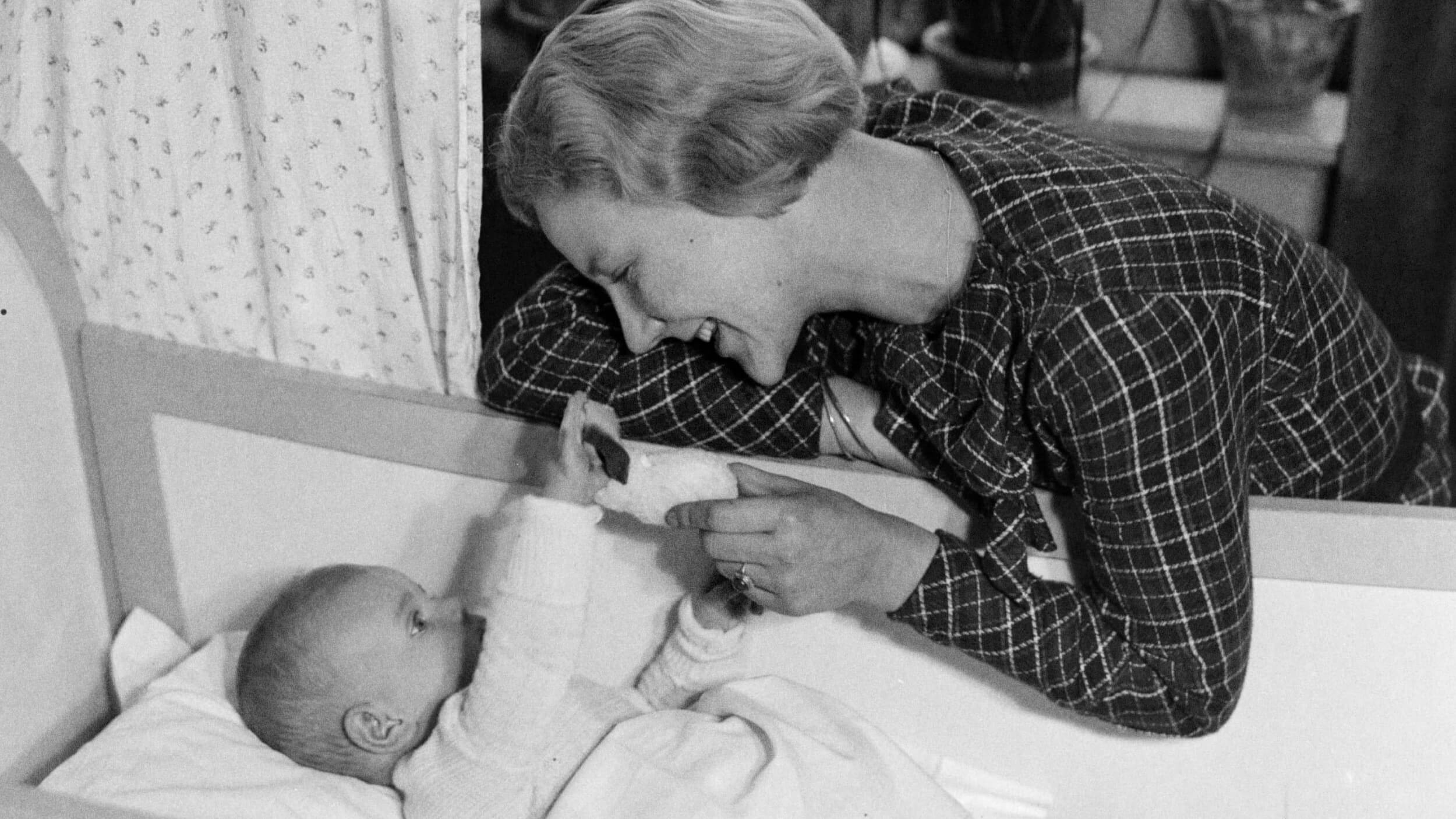 A woman smiles at a baby in a crib, holding a toy while the baby reaches out, attempting their first words. They appear to be in a domestic setting.