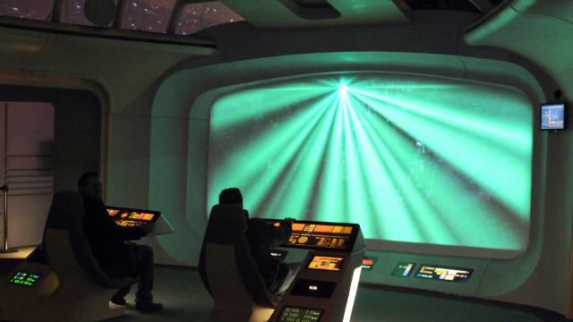 In a futuristic control room, two people sit at advanced control panels, utilizing AI technology as they face a large screen displaying vibrant green light beams.