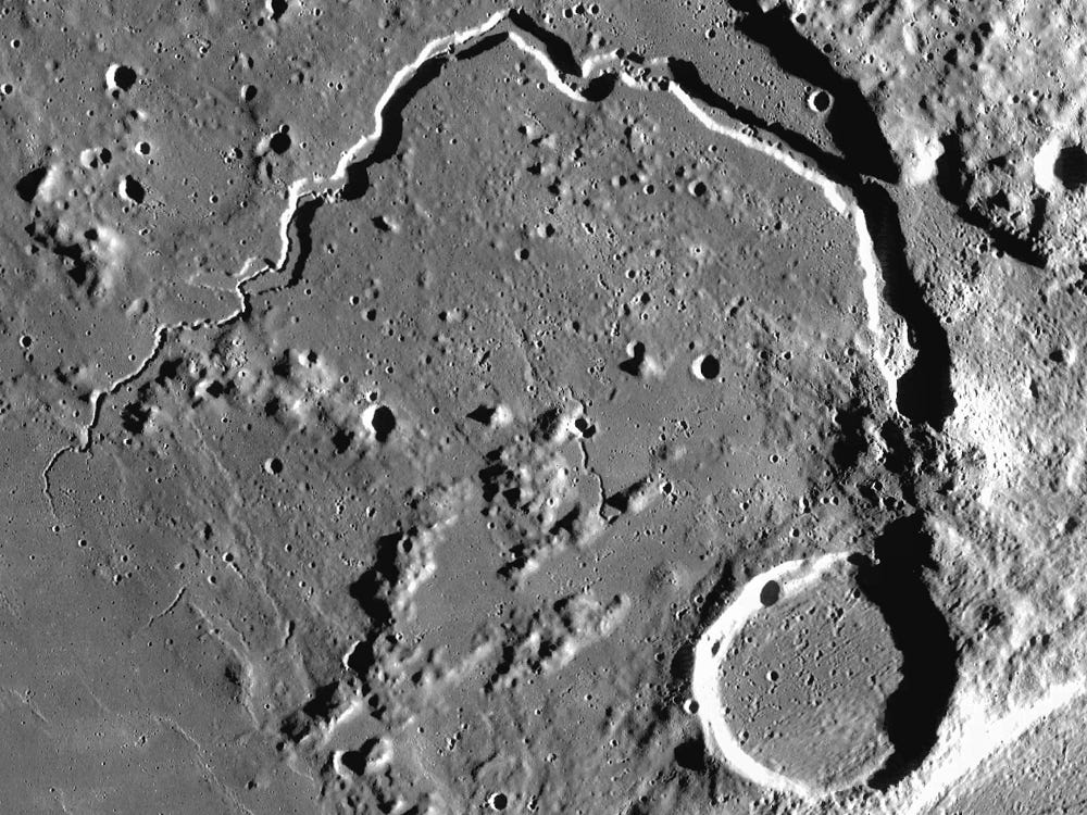 Aerial view of the Moon's surface reveals a winding rille reminiscent of a miniature Grand Canyon and numerous craters, including a prominent circular crater towards the bottom right.