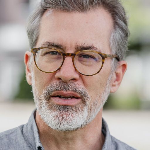 Man with gray hair, beard, and glasses wearing a gray button-up shirt, standing outdoors.