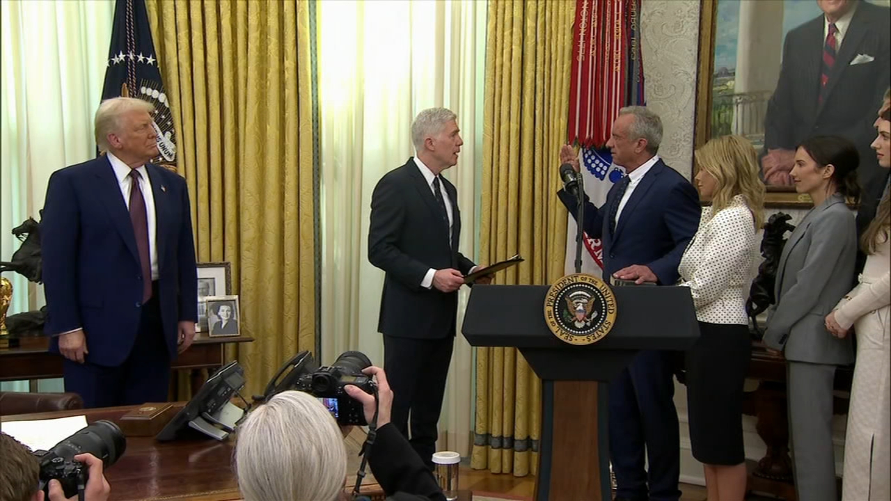 Group of people in a formal setting, with a man holding a large book, others standing nearby, and photographers capturing the scene. There is a large portrait and flags in the background.