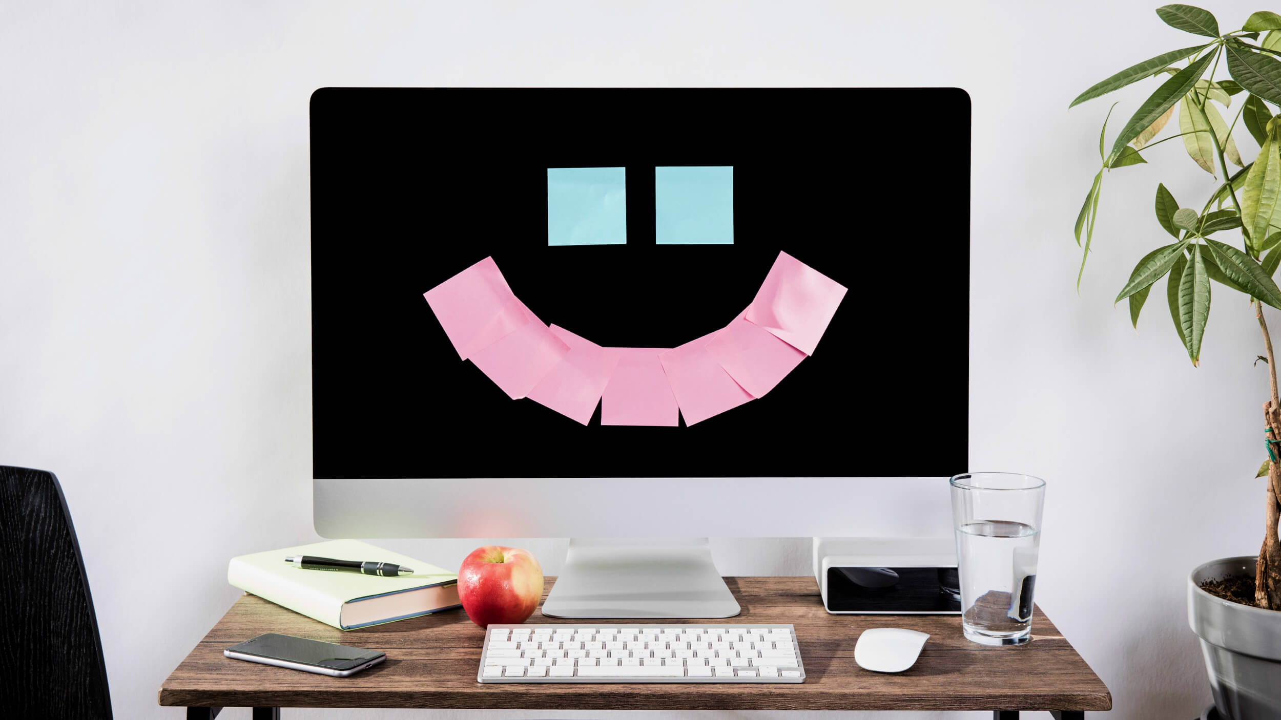 A computer screen adorned with a smiley face made of sticky notes exudes workplace happiness, sitting on a desk alongside a keyboard, mouse, phone, notebook, apple, glass of water, and small plant.