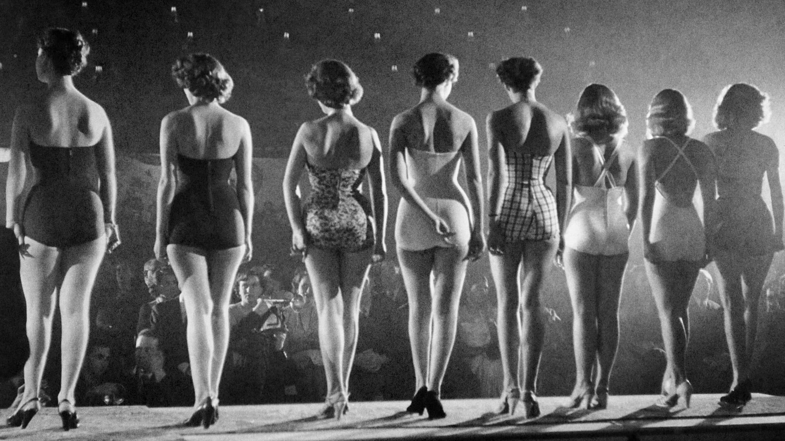 A lineup of eight women in swimsuits stands on stage, embodying beauty as they face away from the camera, with an audience in the background.