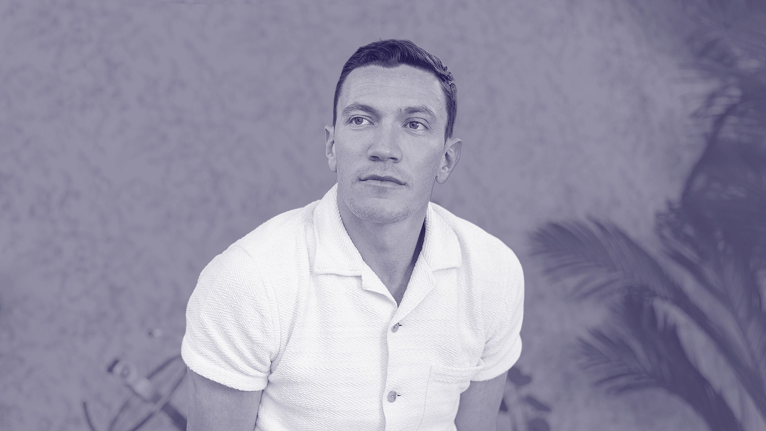 David Perell, in a short-sleeve white shirt, sits in front of a blurred background with plants, looking slightly to the side.