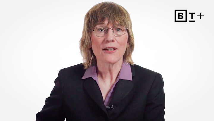 A person with medium-length light brown hair, glasses, and a suit jacket over a purple shirt speaks in front of a white background with a "B T +" logo in the top right corner.