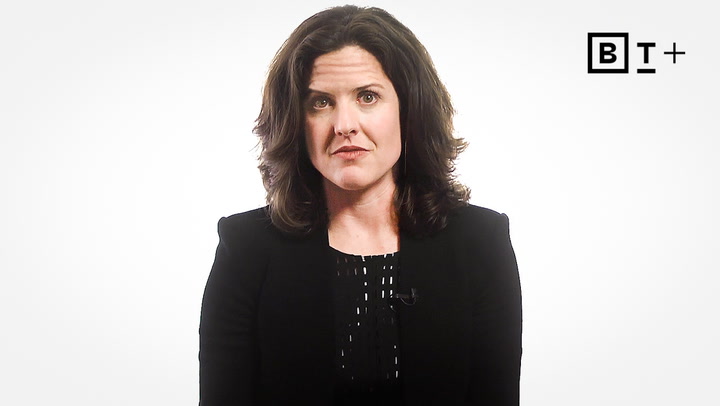 A woman with shoulder-length brown hair, wearing a black blazer and patterned blouse, sits against a plain white background with a "BT+" logo in the upper right corner.