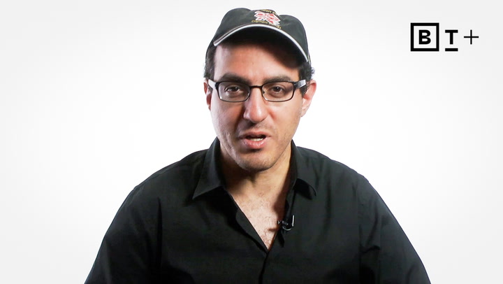 A man wearing glasses, a black shirt, and a black cap is speaking in front of a white background with a "BT+" logo in the top right corner.