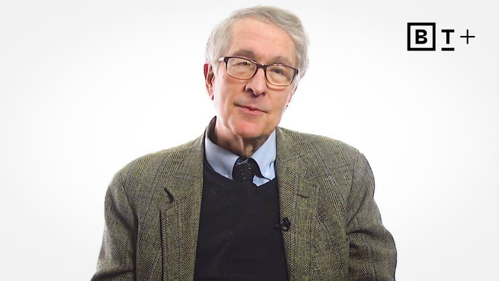 An older man wearing glasses, a brown tweed jacket, and a blue shirt sits against a plain white background with a "B T +" logo in the top right corner.