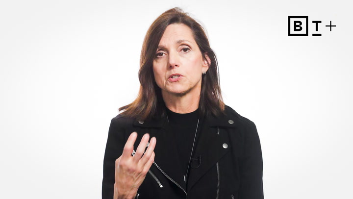 A woman with shoulder-length brown hair, wearing a black jacket, gestures with her hand while speaking against a white background with a "B T +" logo.