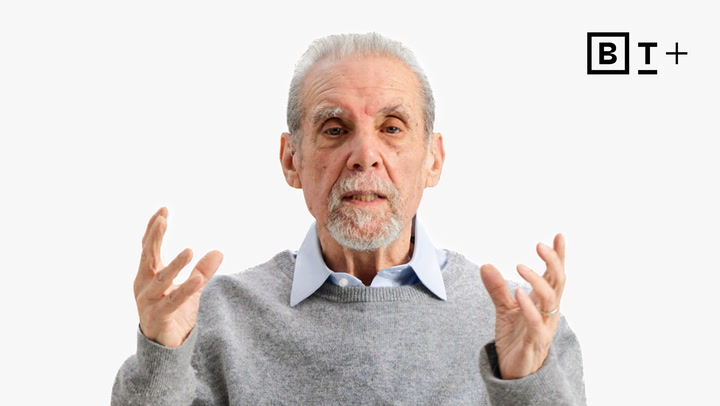 An older man with gray hair and a beard, wearing a light blue shirt and gray sweater, gestures with his hands. The "B T +" logo appears in the top right corner.
