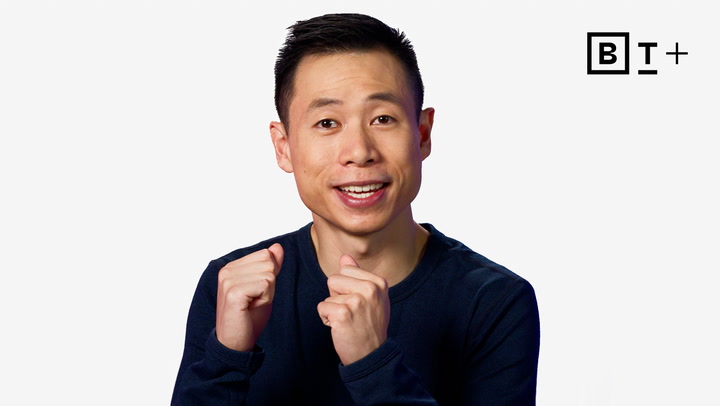 A person in a dark long-sleeve shirt gestures with closed fists and smiles, standing in front of a plain white background with a "B T +" logo in the top right corner.