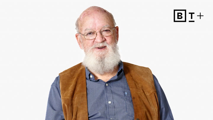 An older man with a white beard and glasses, wearing a blue shirt and brown vest, stands in front of a plain white background with a "BT+" logo in the top right corner.