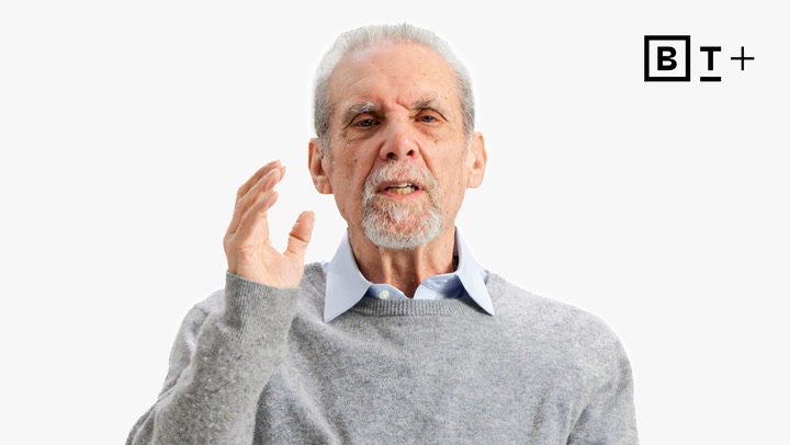 An older man with gray hair and a beard, wearing a gray sweater over a blue collared shirt, gestures with one hand. The letters "B T +" appear in the top right corner.