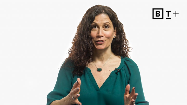 A woman with curly dark hair and a green blouse speaks, gesturing with her hands. The letters "B T +" are visible in the top right corner on a white background.