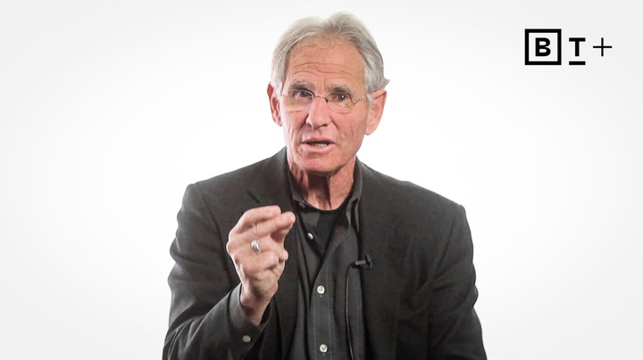An older man in a dark suit and glasses gestures while speaking in front of a plain white background with a "BT+" logo in the top right corner.