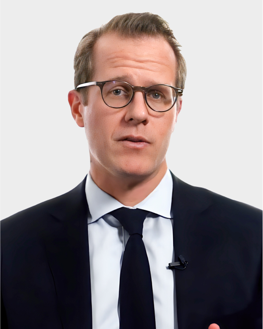 A man wearing glasses, a dark suit, white shirt, and dark tie stands against a plain light background, looking directly at the camera.