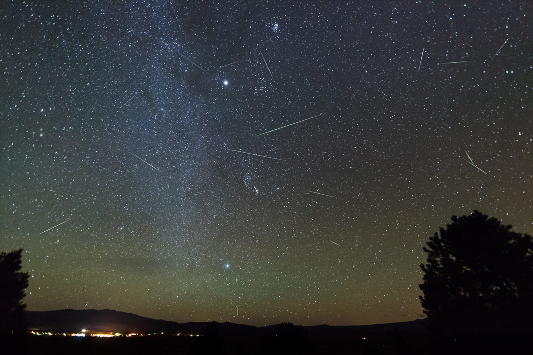 Night sky adorned with countless stars and shooting stars; meteor streaks illuminate the darkness, while the faint silhouette of trees and distant town lights frame the horizon&mdash;perhaps a glimpse of Halley's Comet graces the view.