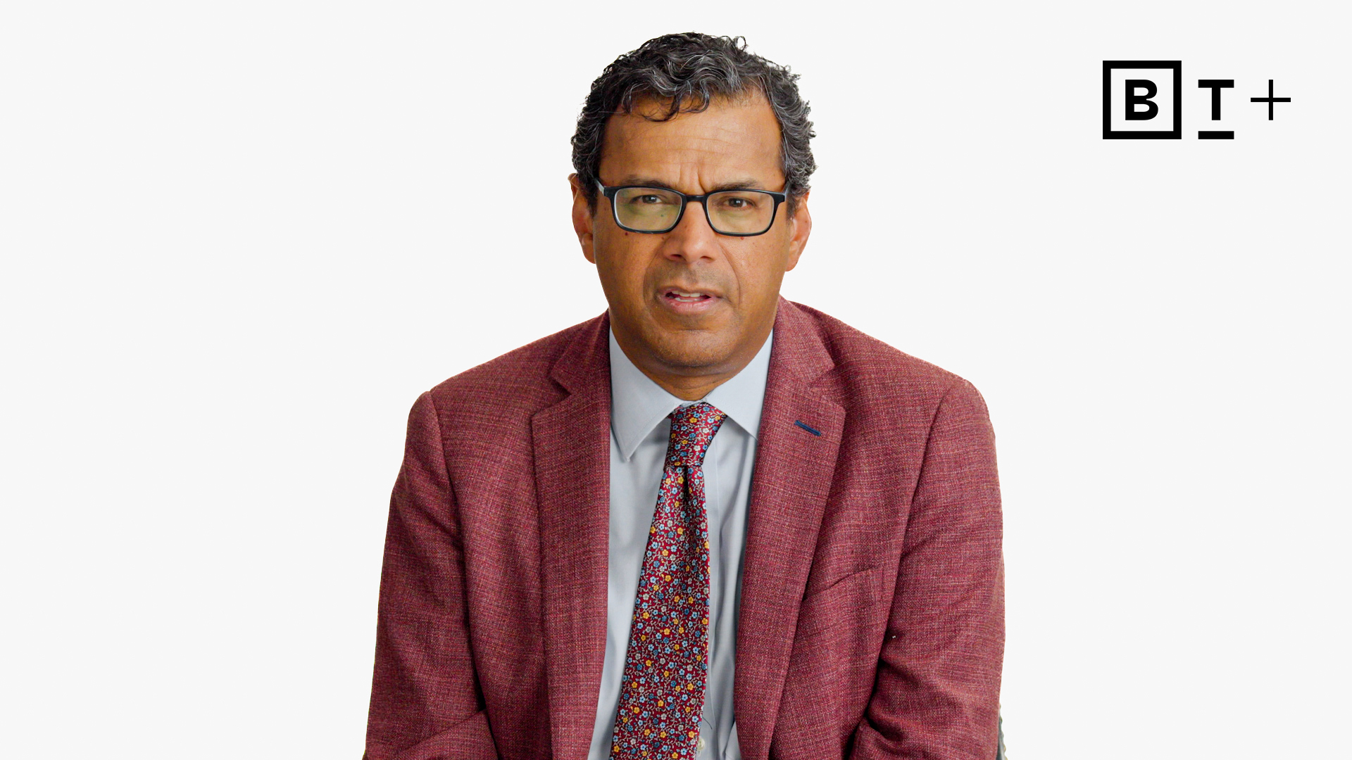 A man in a maroon suit jacket, patterned tie, and glasses sits facing forward against a plain white background with the B+ logo in the top right corner.