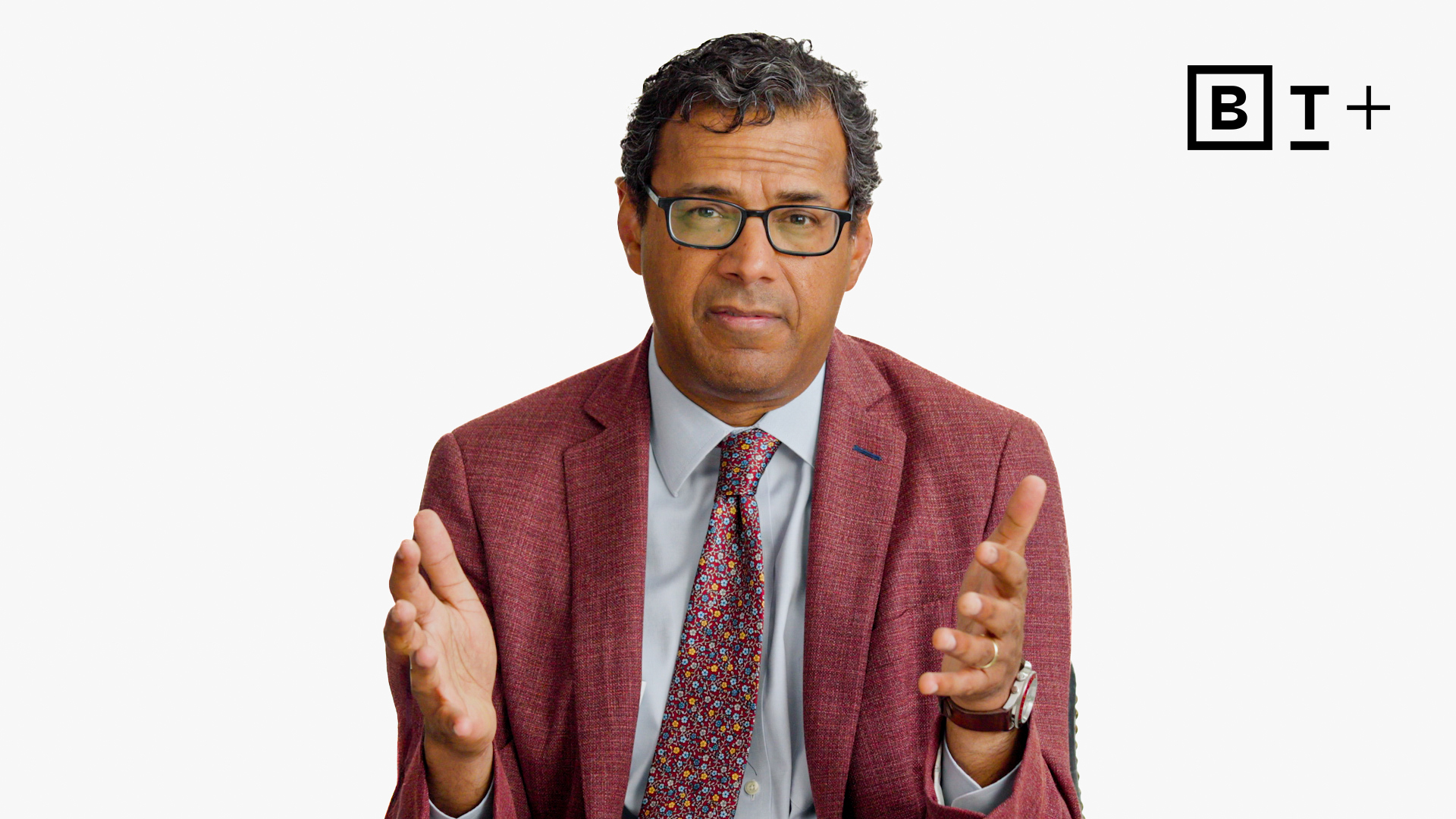 A man in a suit and glasses speaks, gesturing with both hands, seated against a plain white background with a "B T +" logo in the corner.