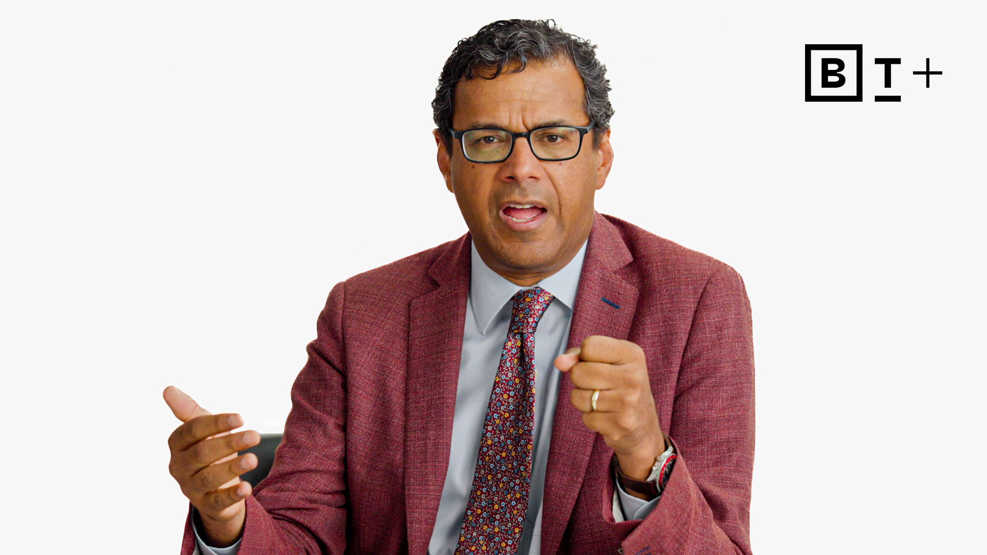 A man in a red suit jacket, dress shirt, and patterned tie speaks with expressive hand gestures in front of a plain white background with the B T+ logo in the top right corner.