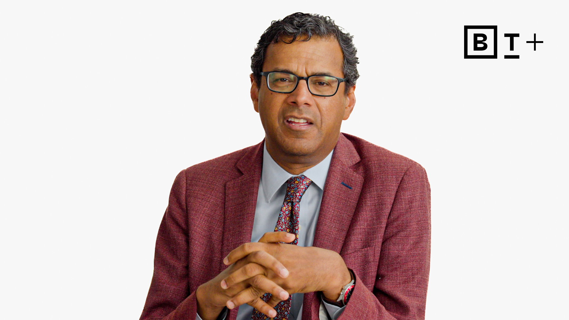 A man in a red jacket and glasses sits against a plain white background, with the "B T +" logo in the top right corner.