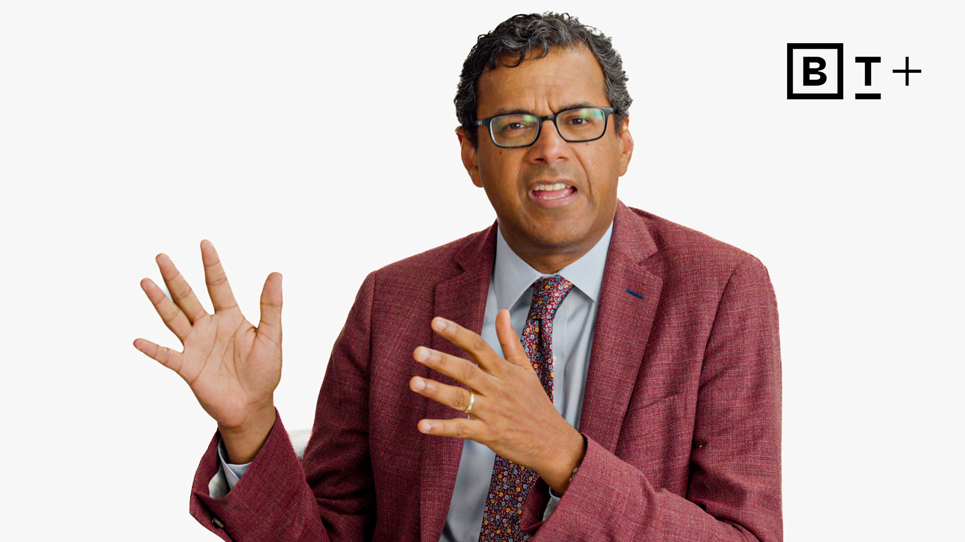 A man in a red suit jacket and tie gestures with his hands while speaking against a white background with a "BT+" logo in the top right corner.